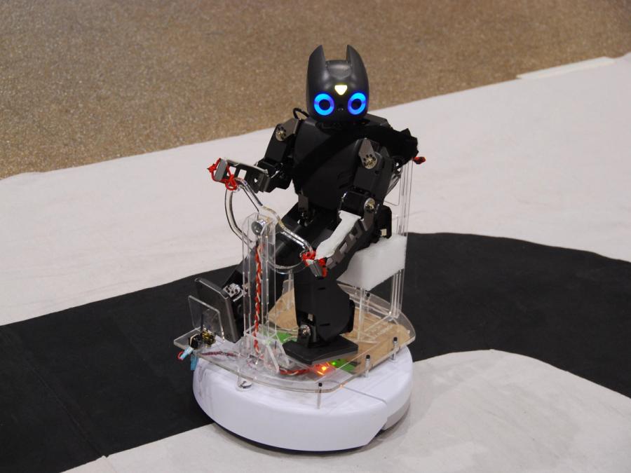 A small humanoid robot appears to be driving on top of a Roomba vacuum.