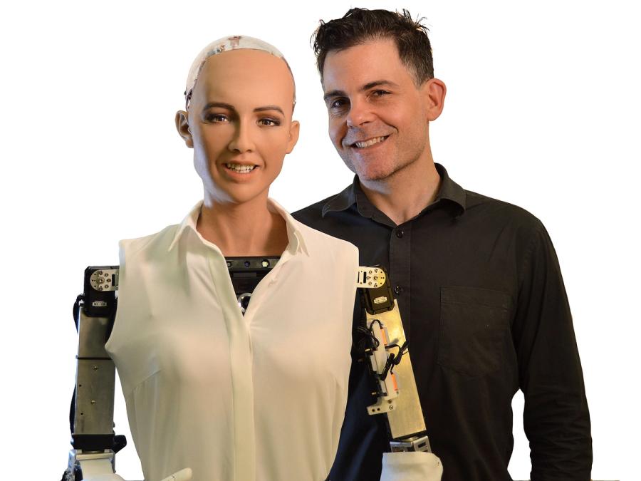 David Hanson, a smiling dark haired man, poses with Sophia, a realistic female humanoid robot whose skin and face mimic a humans.