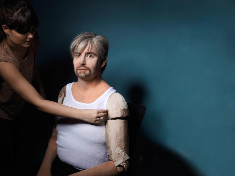 A woman adjusts the arm of a humanoid male robot. The robot is very realistic, other then it's forearm which is exposed and is sectioned in several pieces.