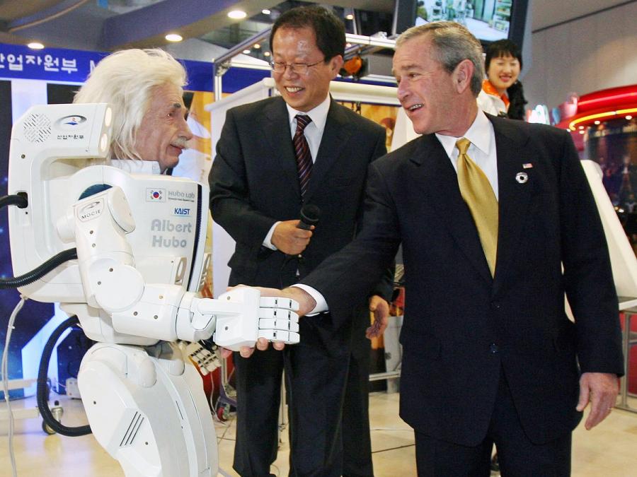 A male humanoid robot shakes the hand of a suited former President Bush while another man looks on.