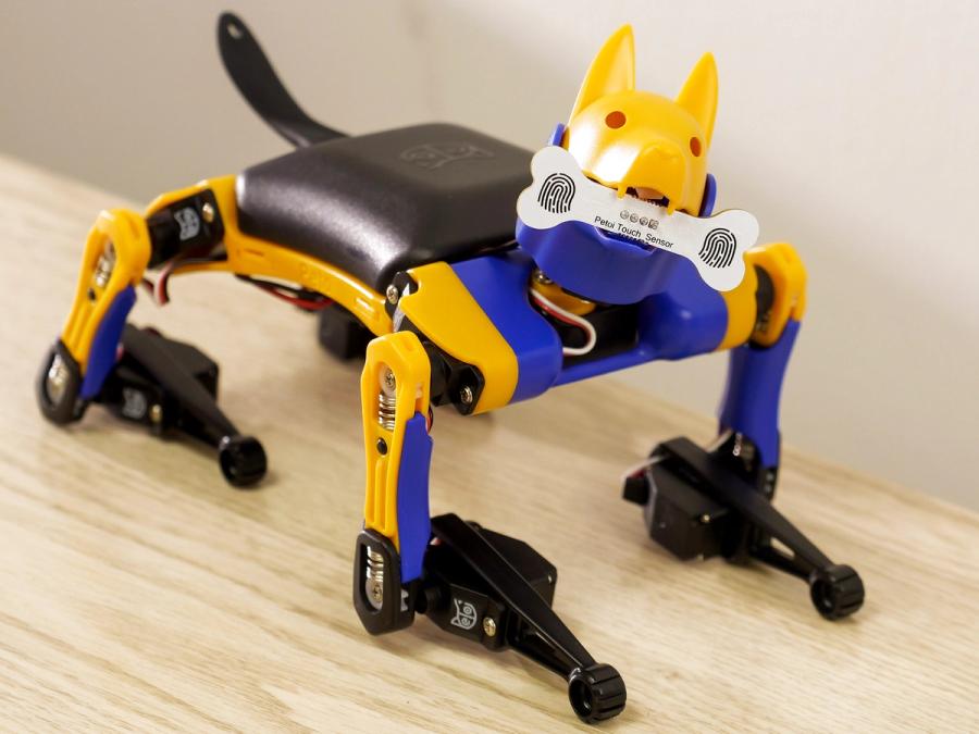 A miniature robot dog holds a wooden bone between it's teeth.