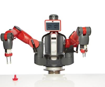 A large red and black robot with two industrial arms with grippers, and a display for a face is placed at a tabletop where it moves its arm and picks up a red object on the table.