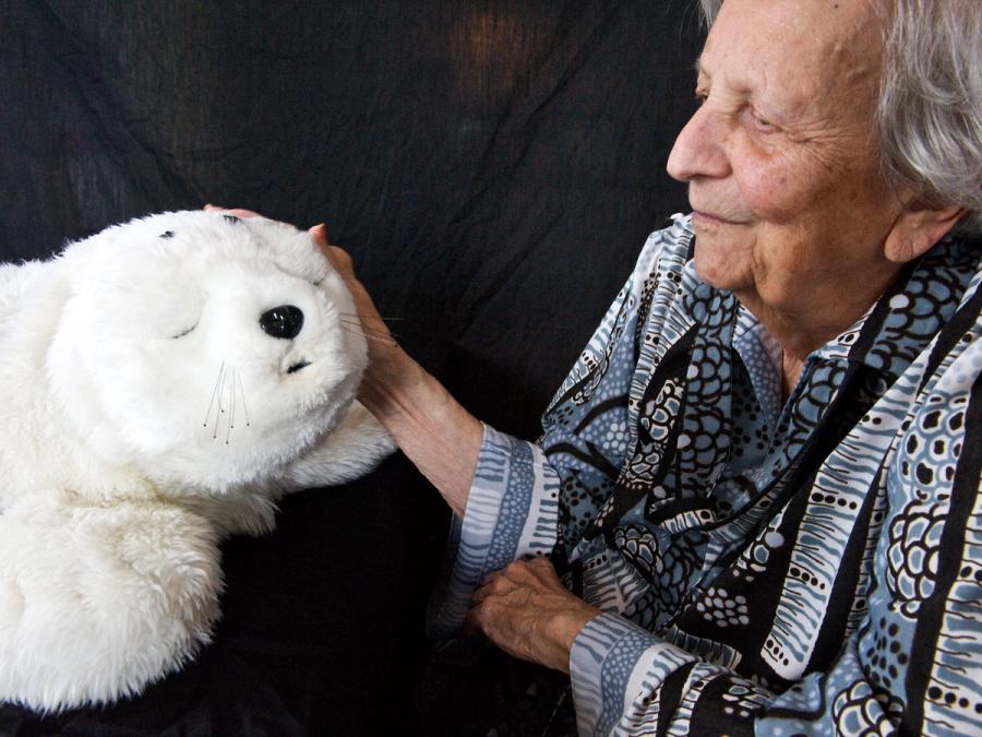 An elderly woman smiles as she gently touches the furry white seal robot.