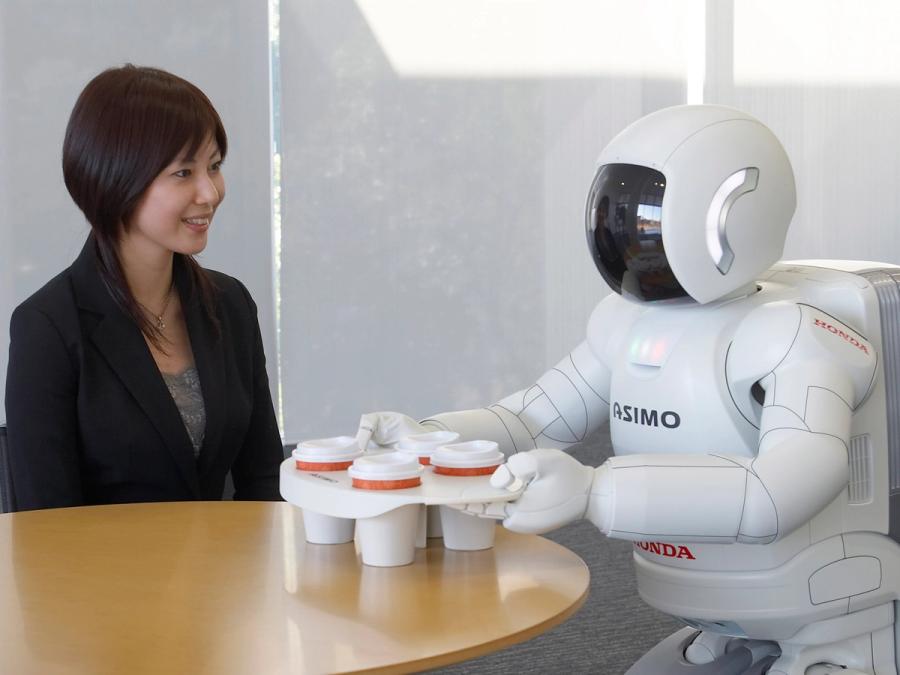 A woman sits at a table while a white robot delivers a holder with four coffee cups.