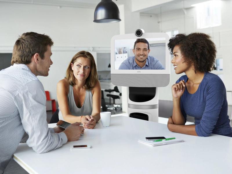 3 people, and a 4th on a telepresence screen, sit around a table.