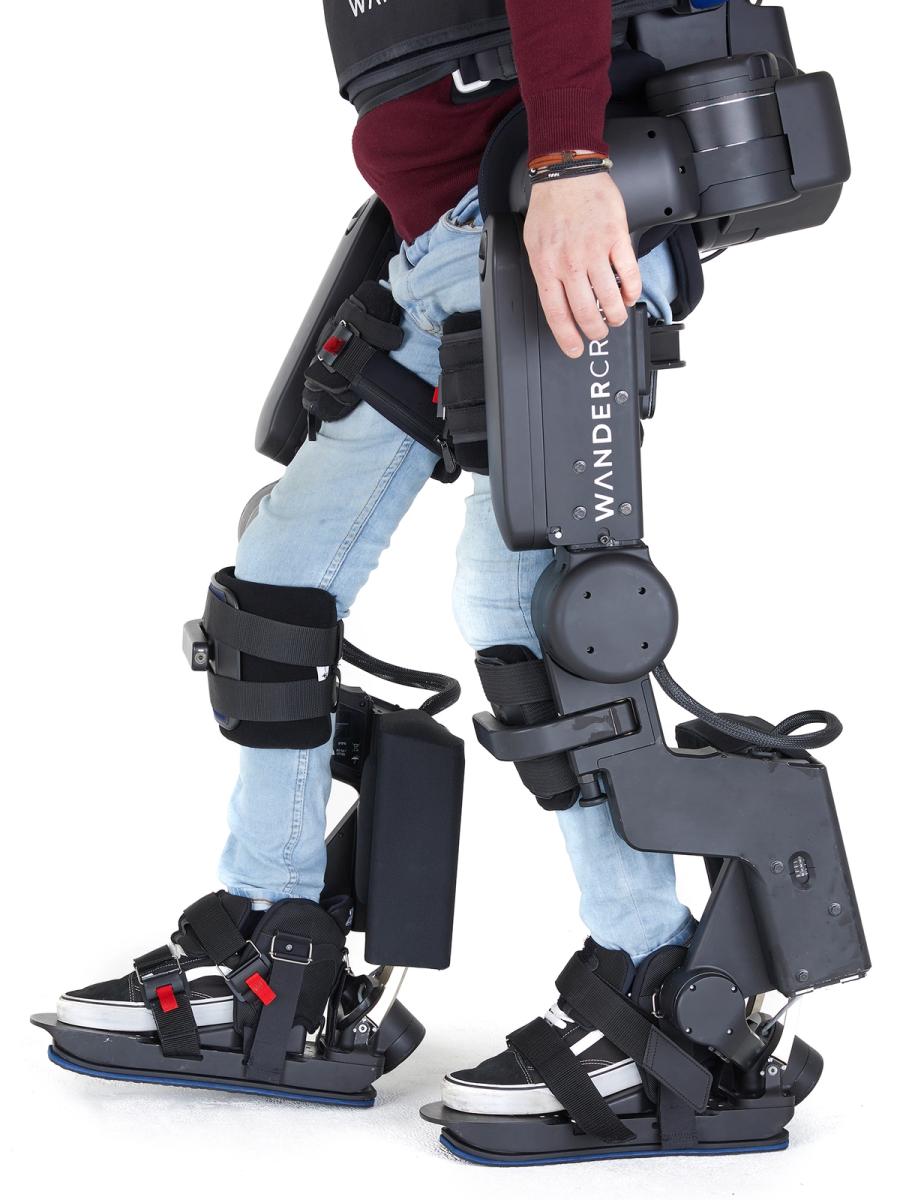 Close-up of a person's legs and the Wandercraft exoskeleton, which includes many black boxes holding motors and equipment, and many straps attaching it to the subject.