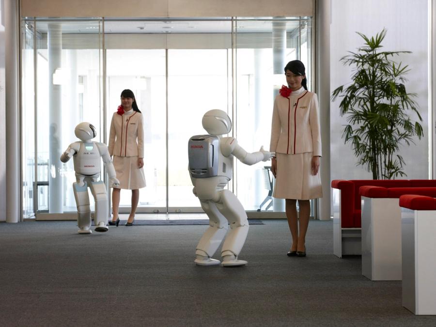 An office with two Japanese women in suits and two white Asimo humanoid robots that are a bit more then half their size.