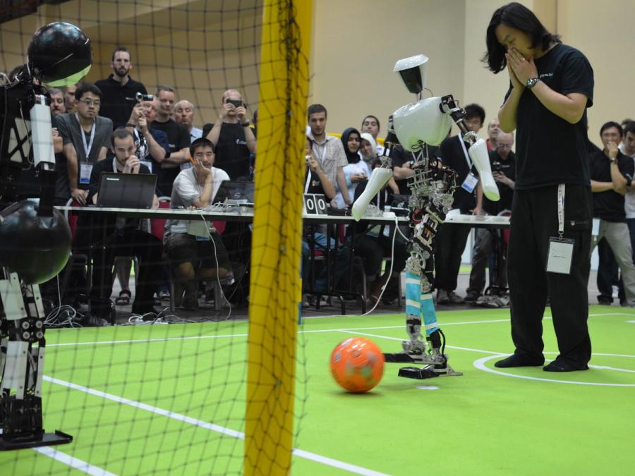 A humanoid robot is almost as tall as the human next to them. They stand on a soccer course surrounded by onlookers as the robot tries to score a goal.