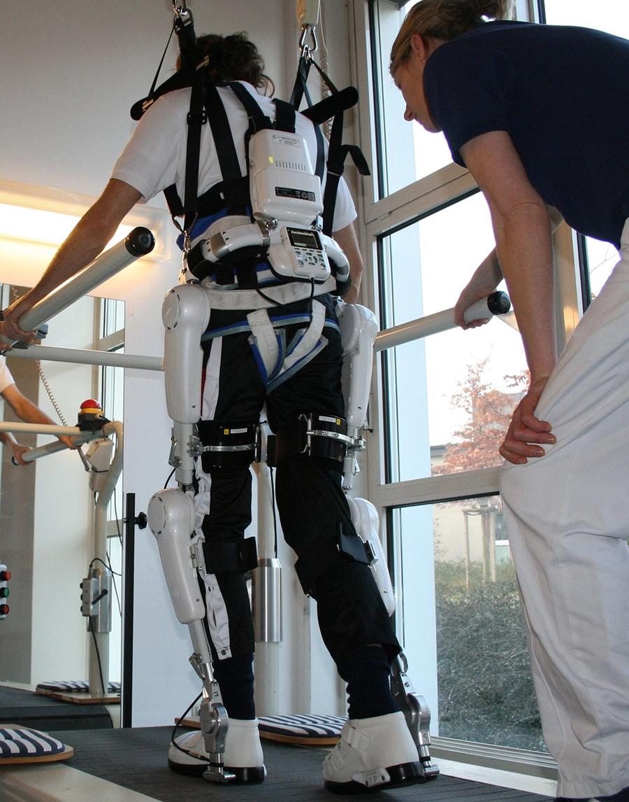 A person on a medical treadmill with rails is attached to a top harness that helps keep them steady in the exoskeleton suit they are wearing. A person stands behind them, watching.