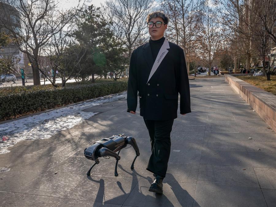 A man in glases and a fancy suit walks down a street with a small quadruped robot by his side.