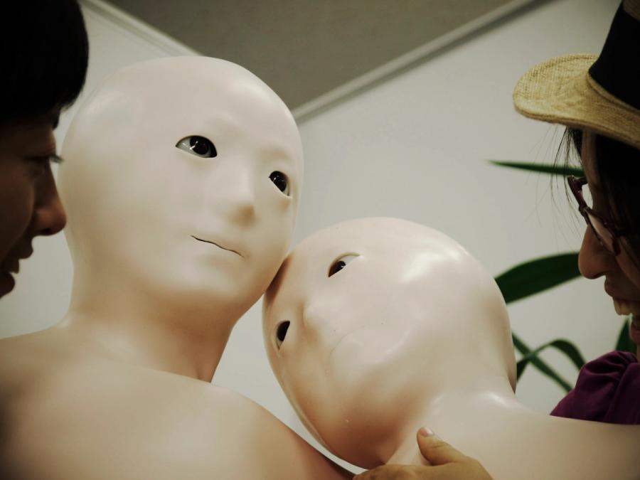 Two robots with soft flesh-colored torsos and doll-like faces are pressed together.