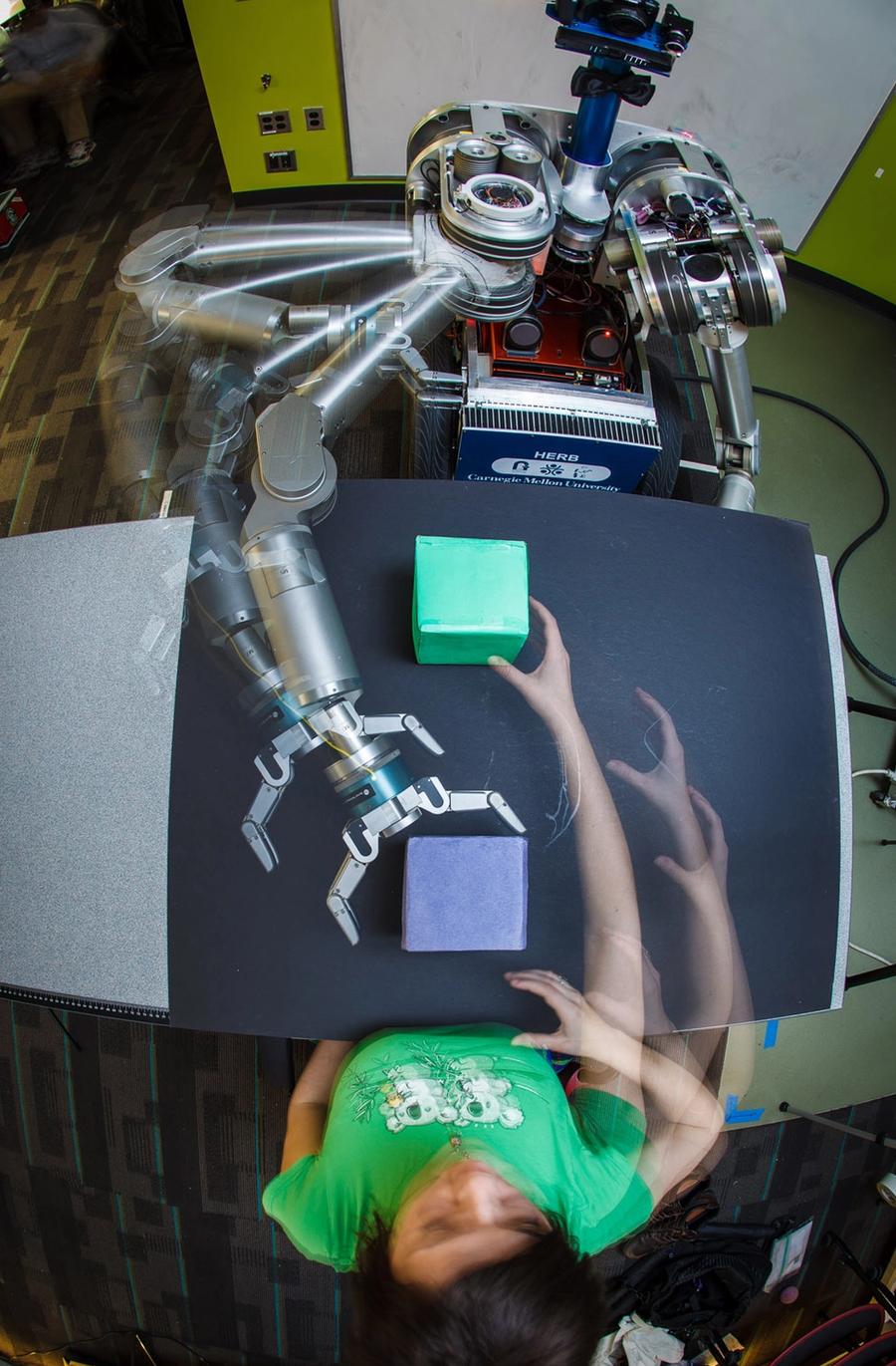 Overhead photograph uses multiple exposures to show the robot and a person sitting across a table from each other. They each reach toward a colored cube in front of each of them, and appear to be mirroring their actions.