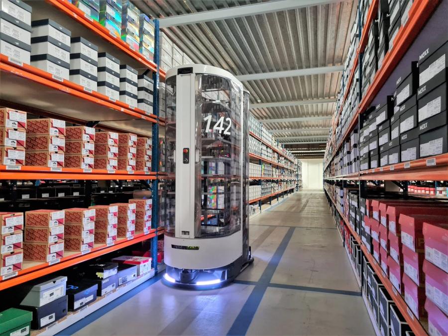 A tall mobile robot with two clear sections joined by a grey based, top and middle section moves on a wheeled, lit base along a warehouse full of shoe boxes on shelves.