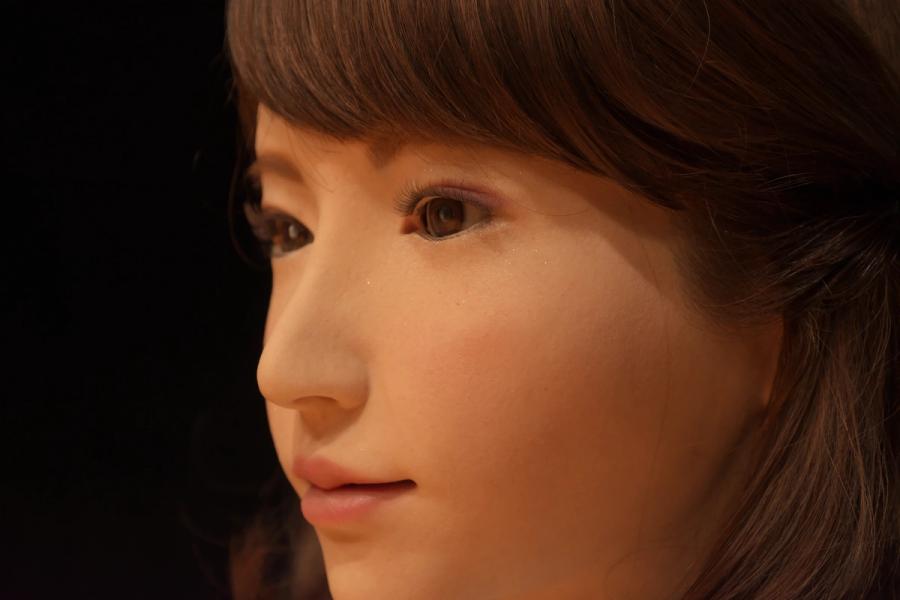 A close-up of female robot Erica's face, which has smooth peach color skin, pink lips, brown almond shaped eyes, and brown hair.