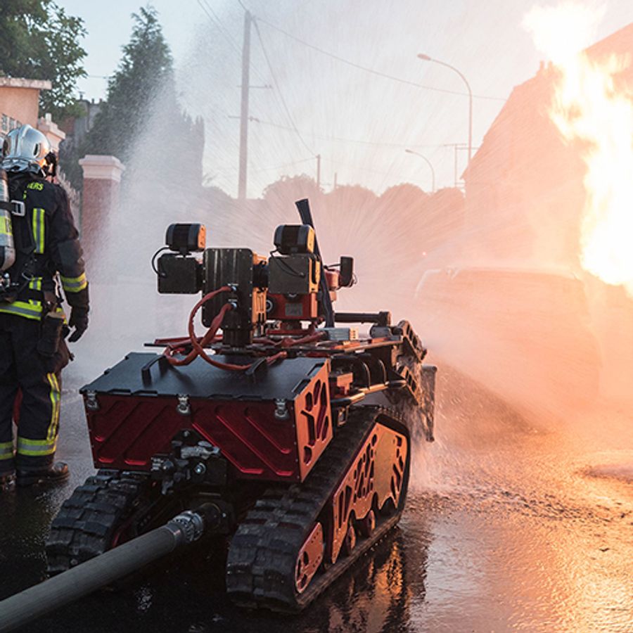 A red robotic vehicle the size of a small tank blasts water on a fire while two human firefighters observe.