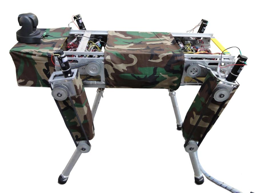 A quadruped robot with a rectangular body (open with wires visible) and four legs has a black camera sitting on top, and is covered in camouflage fabric.