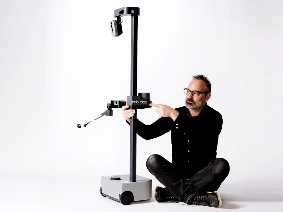 A man in glasses with salt and pepper hair sits crossed legged next to a mobile robot as he adjusts its stretchable arm.