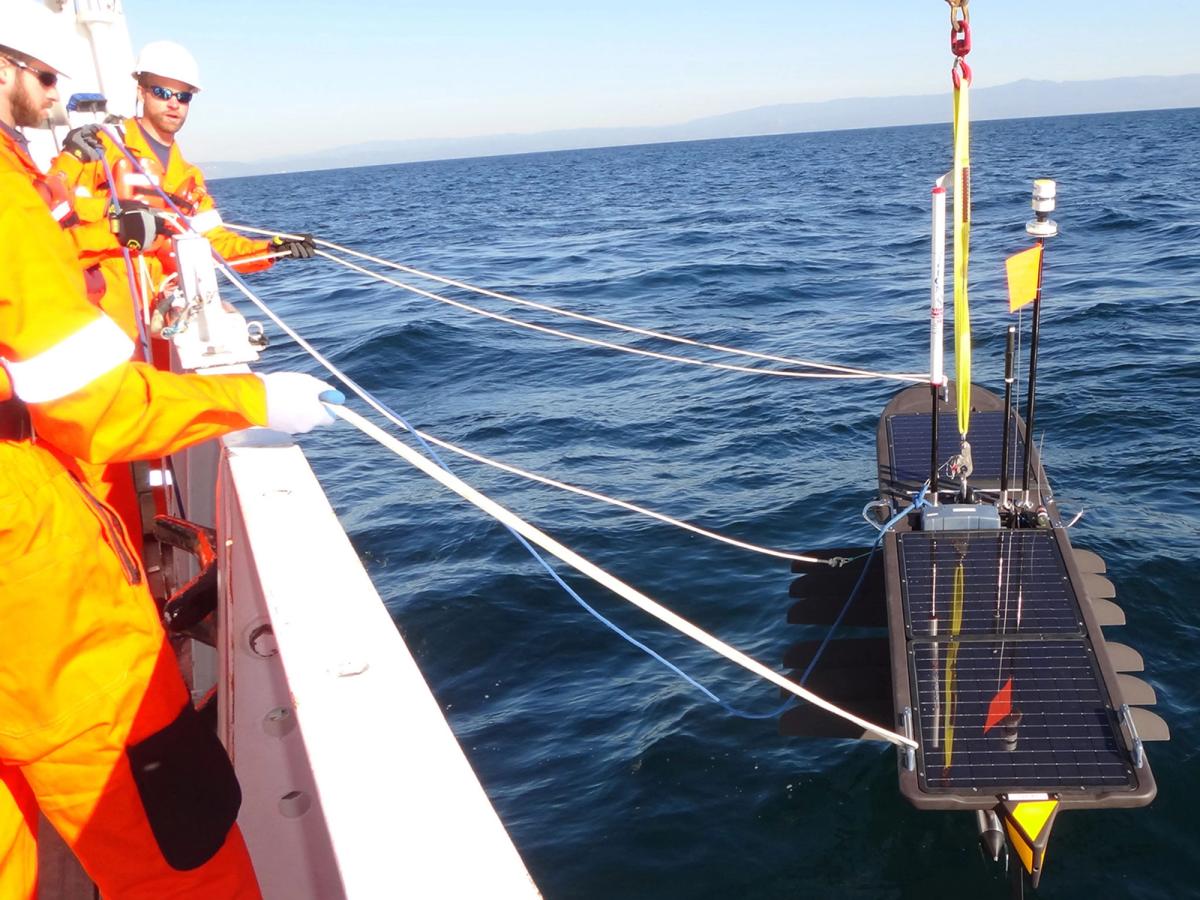 Men in hardhats and orange outfits stand on a boat holding onto white strings that are attached to long board-like robot covered in solar panels and multiple masts, which sits in the water.