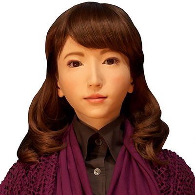 A highly realistic humanoid female robot with golden skin, almond shaped eyes, and shoulder length brown hair wears a black button up shirt and purple sweater.