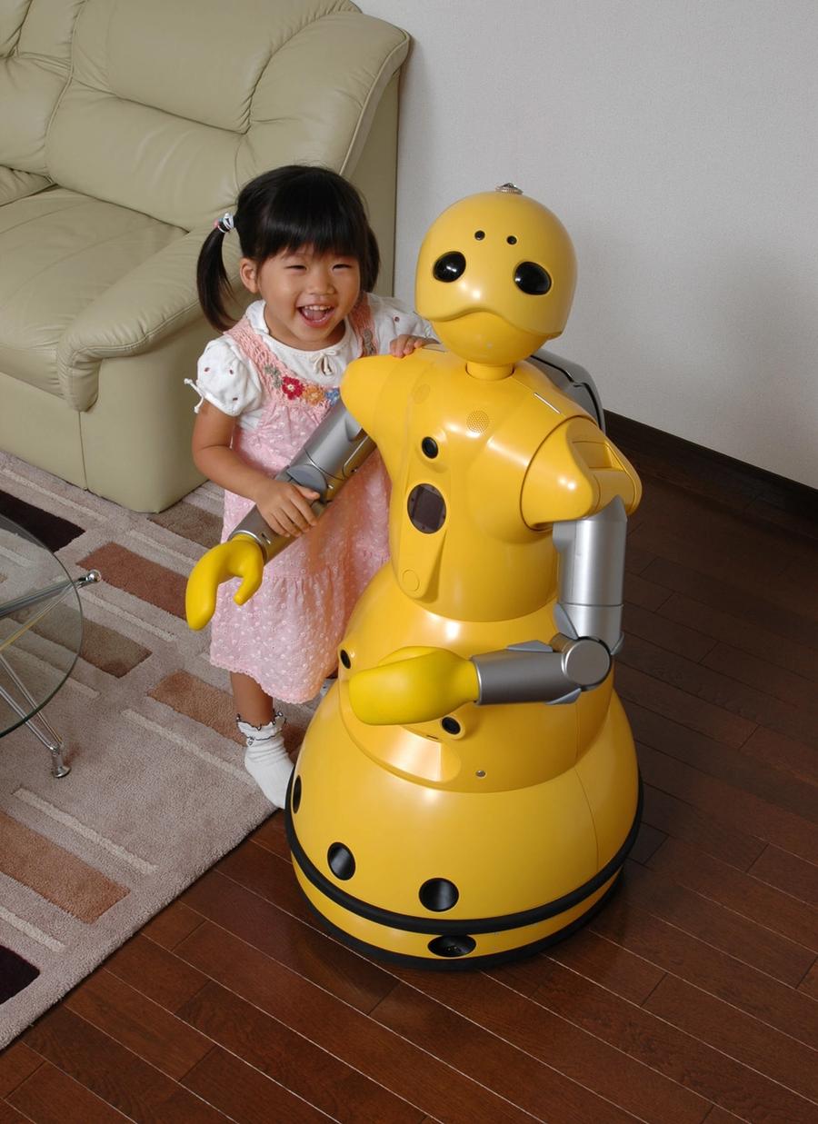 A yellow mobile humanoid robot that is the same size as the smiling toddler holding its arm in a living room.