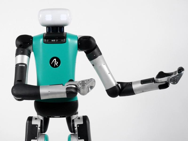 Digit is a teal bipedal robot with long arms, a spherical head with glowing eyes, and many sensors and cameras visible in it's neck. It is holding one arm out to the side. The Agility Robotics logo is on its chest.