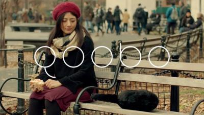 A woman wearing a red beret and scarf sits on a park bench next to Qoobo, a round furry pillow-shaped robot with a tail and no head.