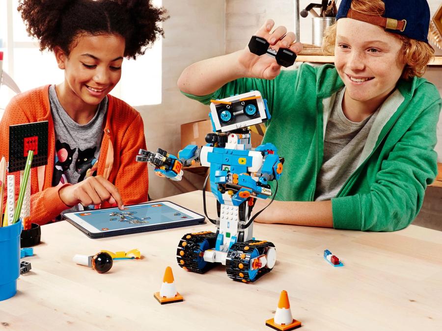Two teenagers use a tablet to program a robot made of Lego bricks forming a humanoid with track wheel feet, two arms, and a cute face with blue eyes and orange eyebrows. 