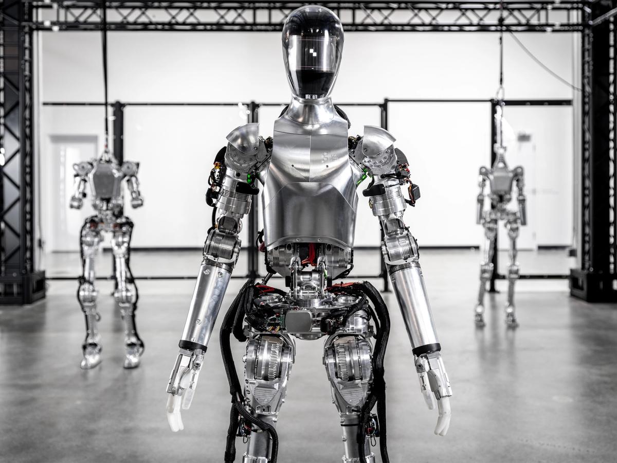 图01机器人，一个闪亮的银色两足类人机器人，双手和头戴头盔，站在仓库里。在它后面是另外两个没有头部的数字机器人。