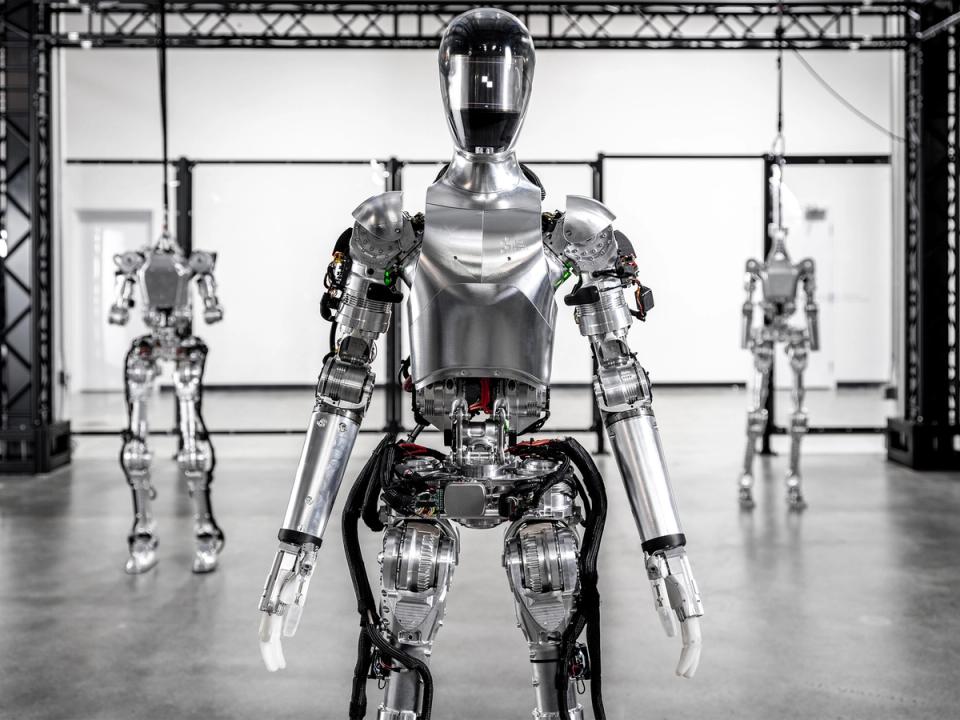 The Figure 01 robot, a shiny silver bipedal humanoid with hands and a helmeted head stands in a warehouse. Behind it are two more Figure robots, but without heads.