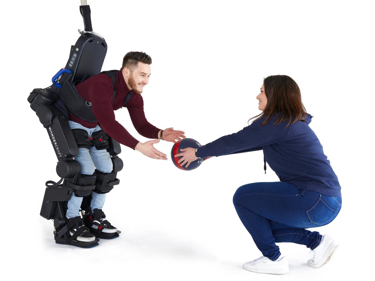 A smiling man wears in an Atalante X exoskeleton that includes strapping around his legs and torso bends down and reaches for a ball that a squatting woman holds out to him.