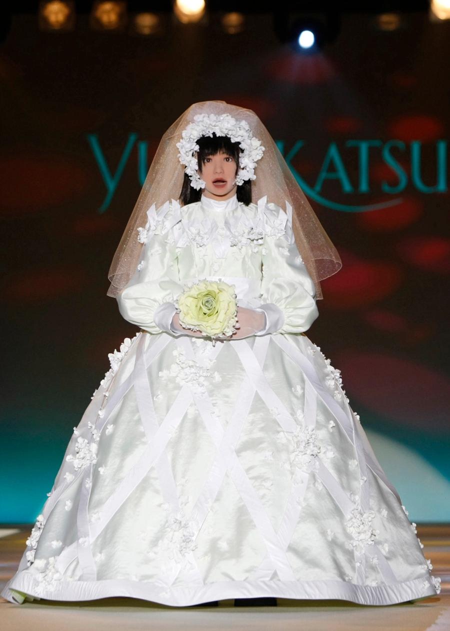 The robot wears an extravagant wedding dress, veil and headpiece. It holds a bouquet of flowers as it walks down a runway.