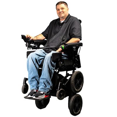 A smiling man clothed in a black shirt and jeans sits in a robotic wheelchair that is elevated on two of its four main wheels.