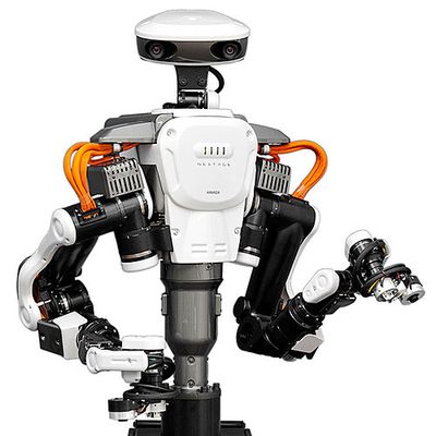 A white, black and orange human sized factory robot with two arms and a head with cameras.