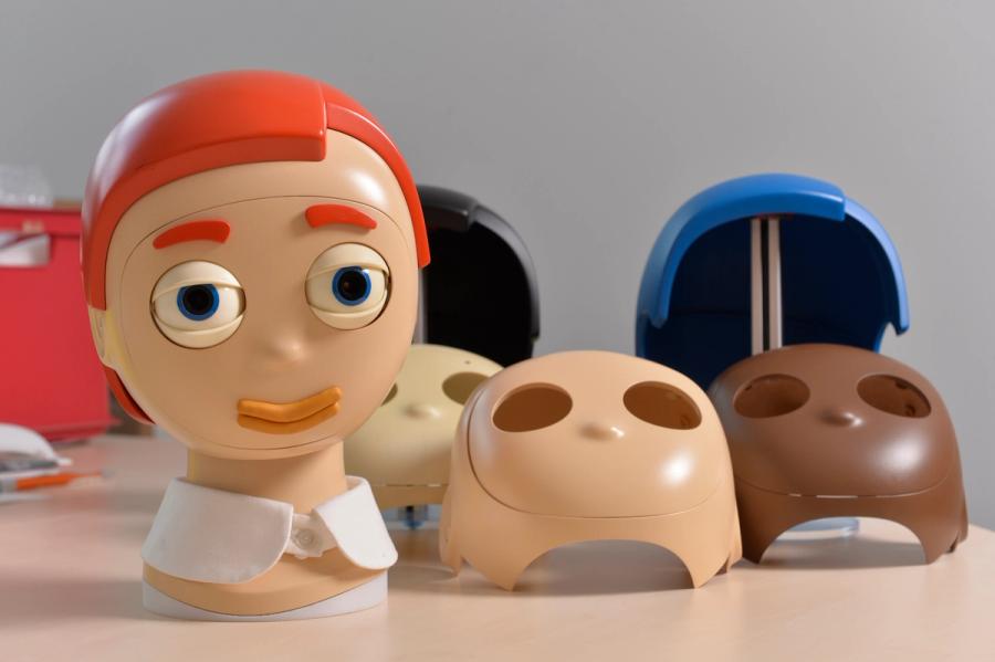 A robotic head with cartoon eyes and orange hair stands on a table next to hair pieces and faces of different colors.