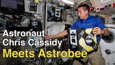 A male astronaut floats inside the space station while gently touching a black and yellow robotic cube that also floats, and superimposed is the phrase Astronaut Chris Cassidy Meets Astrobee.