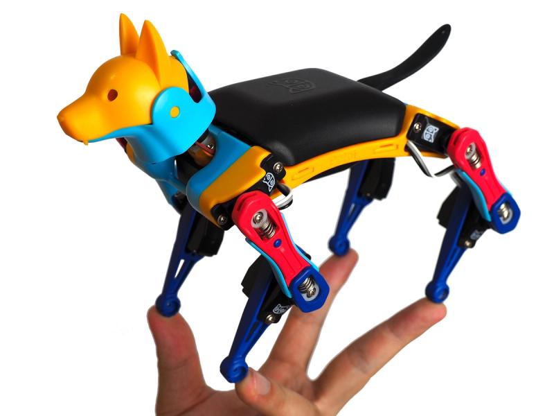 A miniature robot dog with a yellow head, black torso and red and blue neck and limbs sits on 4 fingertips.