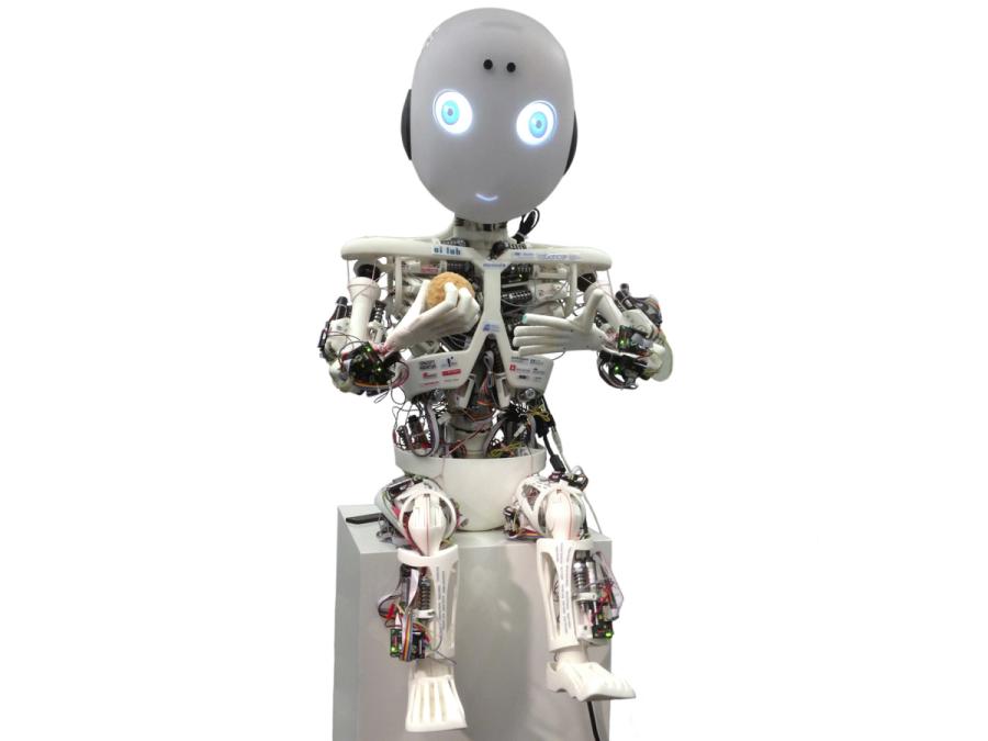 A child-like skeletal robot sits on a block. It's musculature and electronics are exposed. Its head is egg shaped with glowing blue eyes and mouth.  