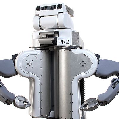 PR2's white plastic and metal torso holds three stacked sections, the bottom which says PR2 and the top of which has two eye cameras and a black bar with sensors. The robots arms, with gripper end effectors, are posed on it's hips.