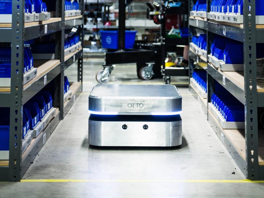 A squat, square, silver mobile robot with two layers and lights works in a warehouse with shelves full of bins.