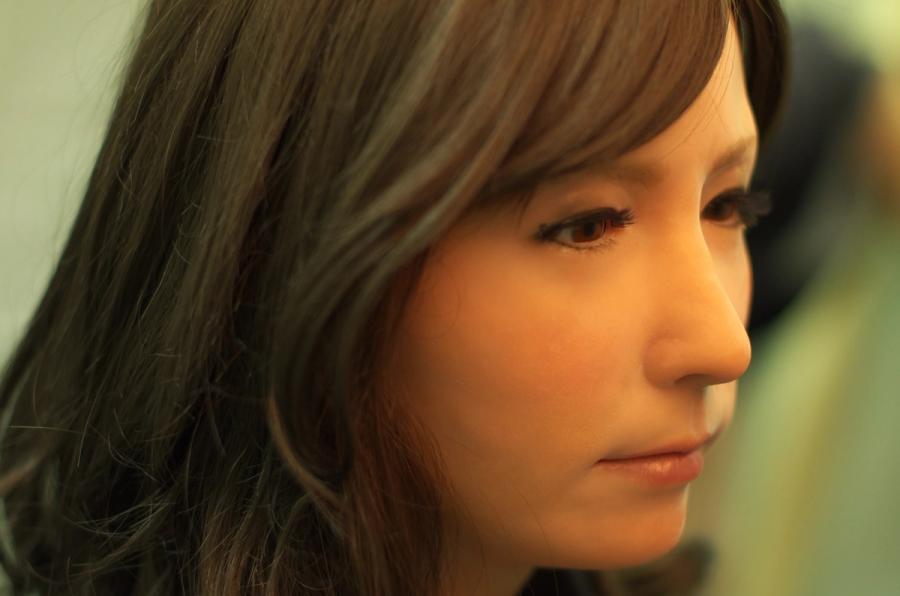 Close-up of female android Geminoid F, with smooth peach color skin, small pink lips, brown eyes and hair.