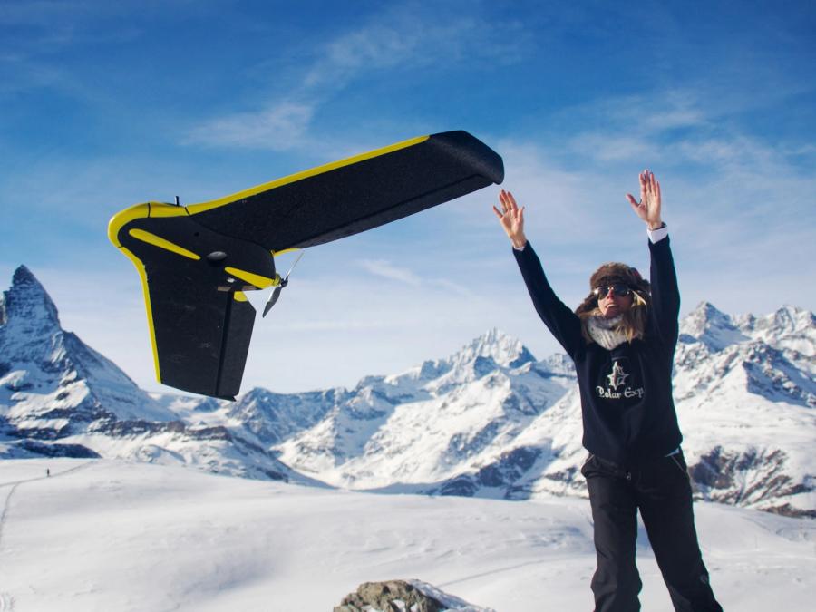 A person in a snowy landscape wearing winter gear tosses a black fixed wing drone with yellow accents and a small spinning propellor.
