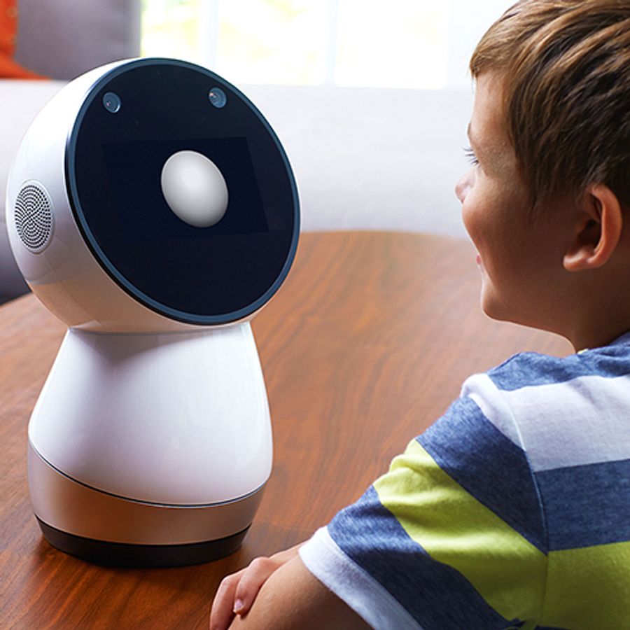 A simple robot with a circular black display screen and bowling pin shaped white and silver base sits on a table and interacts with a young boy.