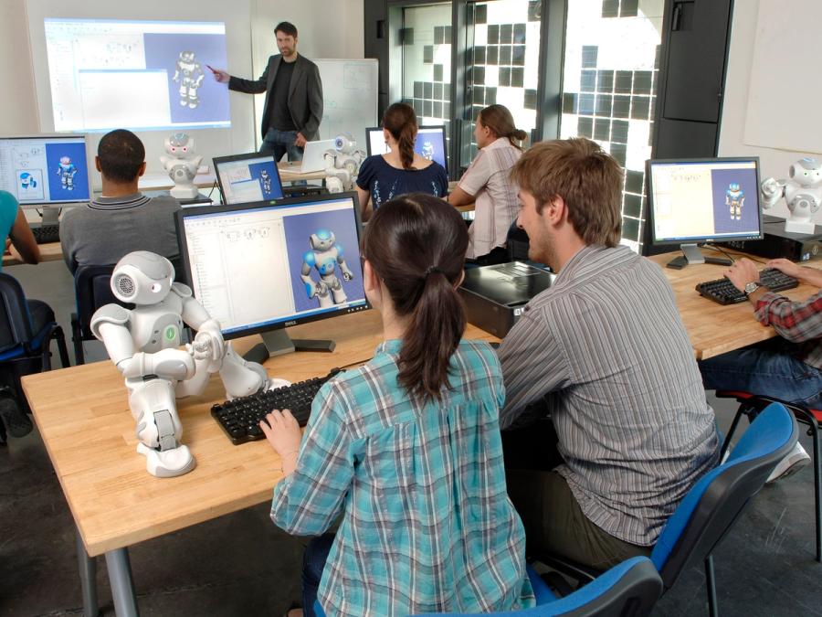 A college computer room with students working at tables with a Nao robot and a monitor showing the work they are doing with the robot. A teacher in the front points at a Nao on the screen.