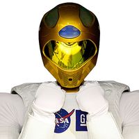 Close-up of Robonaut's gold helmet. It wears cushiony white clothes with the NASA logo on them.