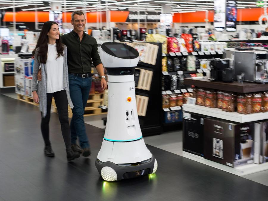 A couple walks behind the wheeled robot in a store.