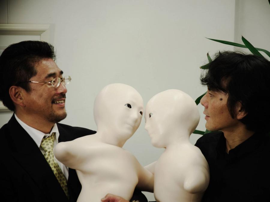 Two robots with soft flesh-colored torsos and doll-like faces are pressed together while held by two men.