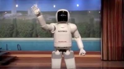 Humanoid robot Asimo, with a white body and astronaut-like head, stands on a stage facing foward and waving its right arm.