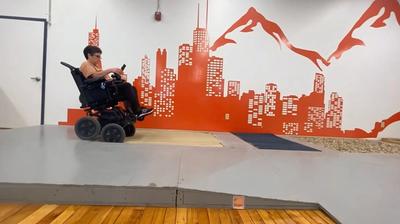 A woman sits on a iBOT powered wheelchair and drives over sand during a test of the machine in a bright room with hardwood floors and white and red walls..