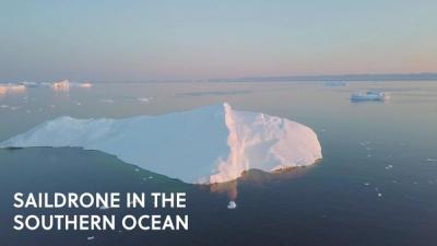 A bright white glacier floats in the Southern Ocean while a small boat sails nearby against a blue and pink cloudless sky.