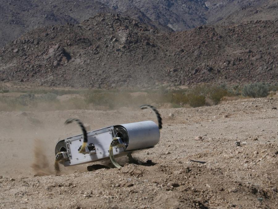 A silver robotic hexapod moves through a rocky desert. It's curved flipper arms are kicking up dirt.